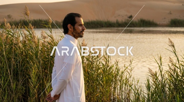 رجل سعودي بالثوب الأبيض عند بحيرة صحراوية وقت الغروب