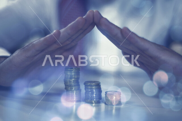 صورة مقربة لرجل عربي خليجي سعودي يضع يديه بشكل هرمي على طاولة فوق مجموعة من العملات النقدية المالية مدموجة بخلفية ضبابية ، الأمن المالي ، تحقيق الثروة المالية ، توفير و إدخار المال ، الإقتصاد و النمو المالي 