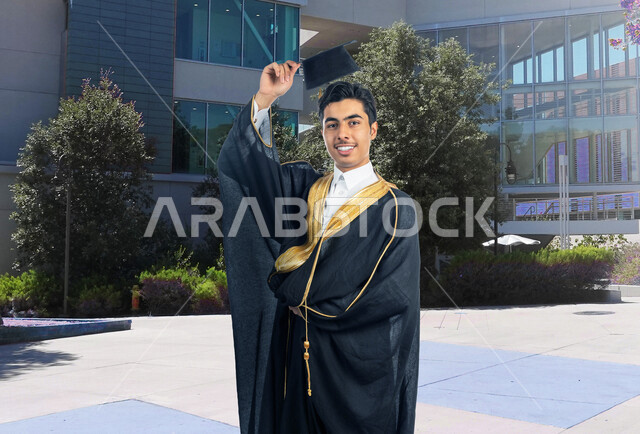 صورة لطالب جامعي عربي خليجي سعودي يرتدي عباءة التخرج، يمسك بيده قبعة التخرج , ايماءات وجه تدل على الفرح والسعادة , حفل تخرج جامعي ، لباس التخرج ، انهاء سنوات الدراسة الجامعية , التخصصات الجامعية .
