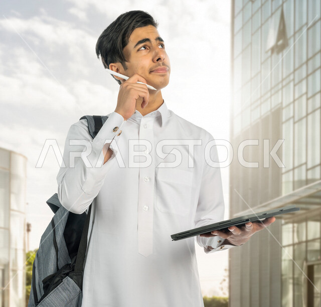 صورة لطالب جامعي عربي خليجي سعودي يرتدي الثوب السعودي التقليدي ، يمسك بيده جهاز التابلت للتعلم ، ايماءات التفكير , طالب هندسي , أفكار انشائية ومعمارية , رؤية مستقبلية ، التعلم عن بعد , الجد والإجتهاد والنجاح . 