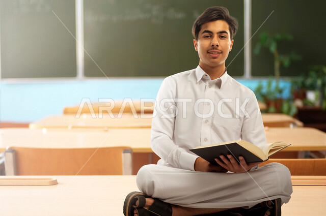 صورة لطالب دراسي عربي خليجي سعودي يرتدي الثوب السعودي التقليدي ، يمسك بيده كتاب جامعي للدراسة ، تأدية الواجبات ، التعلم الذاتي , الجد والإجتهاد , التعليم الأكاديمي , مرحلة دراسية , العودة إلى المدارس , الفصل الجامعي بالخلفية . 