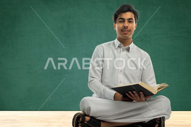 صورة لطالب دراسي عربي خليجي سعودي يرتدي الثوب السعودي التقليدي ، يمسك بيده كتاب جامعي للدراسة ، تأدية الواجبات ، التعلم الذاتي , الجد والإجتهاد , التعليم الأكاديمي , مرحلة دراسية , العودة إلى المدارس ,لوح سبورة خضراء تعليمية بالخلفية . 