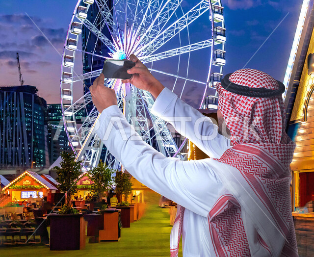 صورة لرجل عربي خليجي سعودي يقوم بالتقاط صورة تذكارية من خلال الهاتف المحمول في مدينة الملاهي ، كاميرا امامية ، الاستمتاع بوقت الفراغ , اجازة صيفية , موسم الرياض , ملاهي وترفيه , قضاء وقت ممتع , السياحة في المملكة العربية السعودية .