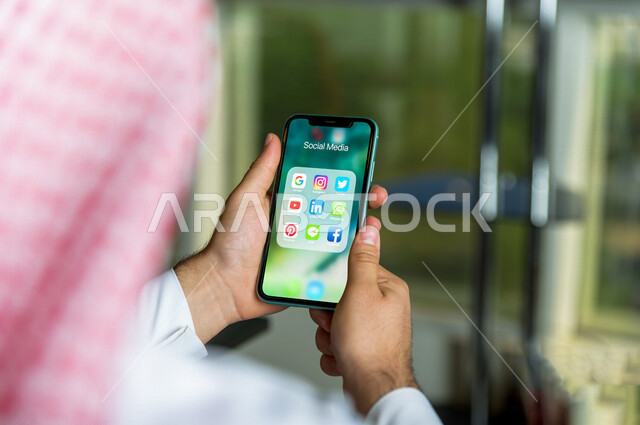 صورة مقربة لرجل عربي خليجي سعودي يقوم بإستخدام هاتفه المحمول ، مجموعة من تطبيقات السوشل ميديا تظهر على شاشة الجوال , تطبيقات التواصل مع الأصدقاء , تصفح مواقع التواصل الإجتماعي , تسلية وترفيه , تعارف وصداقات .