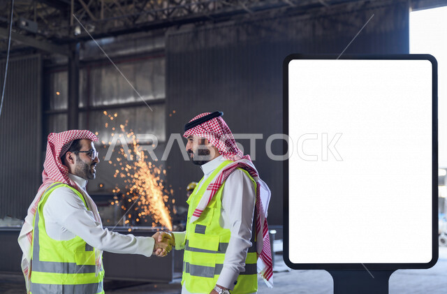 مهندسان عربيان خليجيان سعوديان يرتديان  سترة الوقاية الخاصة بالعمل يتصافحان بسعادة لبدء مشروع جديد ، نجاح العملية الإنتاجية ، لوحة يونيبول إعلانية تجارية مضيئة بيضاء ، إعلانات دعائية و تسويقية ، موك أب لوحة إعلانية 