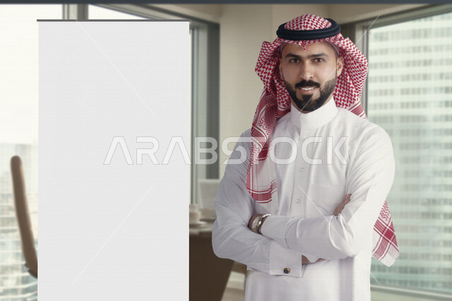 رجل عربي خليجي سعودي مبتسم يقف بجانب لوحة ينيبول إعلانية تجارية بيضاء في الشركة ، عقد صفقات تجارية إعلانية ، لوحة إعلانية فنية تجارية ، عروض تسويقية ، دعاية و إعلان ، موك أب لوحة إعلانية 