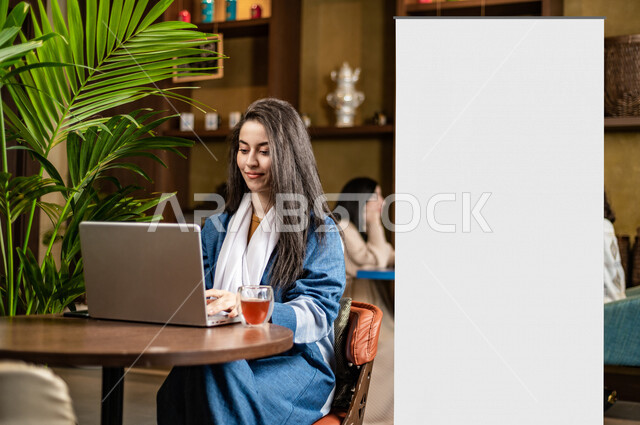 A young Saudi Arabian Gulf woman using her laptop sitting next to a white illuminated commercial unipol billboard in a coffee shop, advertising business deals, commercial art billboard, promotional and marketing offers, billboard mockup