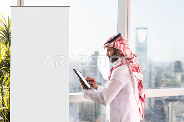 رجل عربي خليجي سعودي يحمل بيده جهاز تقني (تابلت ) يقف بجانب لوحة يونيبول إعلانية تجارية مضيئة بيضاء في الشركة ، عقد صفقات تجارية إعلانية ، إدارة العمل عن بعد ، التسويق الإلكتروني ، موك أب لوحة إعلانية 