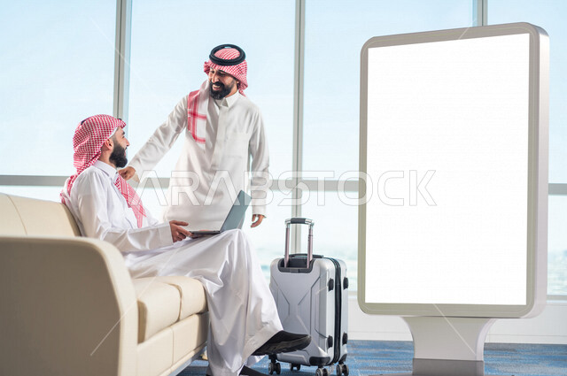 رجل عربي خليجي سعودي بيده حاسوب محمول يتبادل الحديث مع رجل سعودي بإيماءات تدل على السعادة ، يجلس في قاعة الانتظار بجانب حقيبة سفر و لوحة يونيبول إعلانية تجارية مضيئة بيضاء ، تسويق شركات الطيران ، حجز تذاكر الطيران ، موك أب لوحة إعلانية 