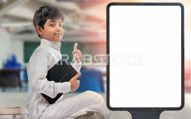 تلميذ مدرسي عربي خليجي سعودي مبتسم يجلس في غرفة صفية يحمل بيده كتاب و يشير بأصبع الابهام لأعلى للرضا و العمل الممتاز ،  لوحة يونيبول إعلانية تجارية مضيئة بيضاء ، إعلانات دعائية تعليمية ، موك أب لوحة إعلانية 