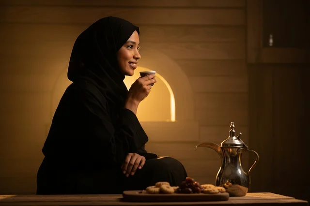 A Saudi woman in an abaya is drinking Arabic coffee next to the oven.