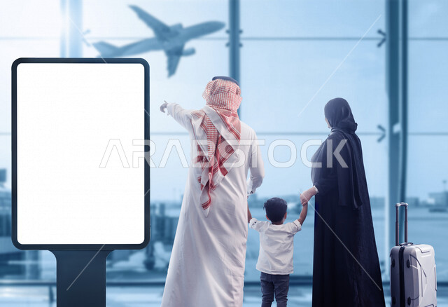 صورة من الخلف لعائلة عربية خليجية تقف بجانب لوحة يونيبول إعلانية تجارية مضيئة بيضاء في قاعة الانتظار في المطار تقوم بمشاهدة إقلاع الطائرة ، رحلة عائلية سياحية ، إعلانات دعائية و تسويقية ، موك أب لوحة إعلانية 