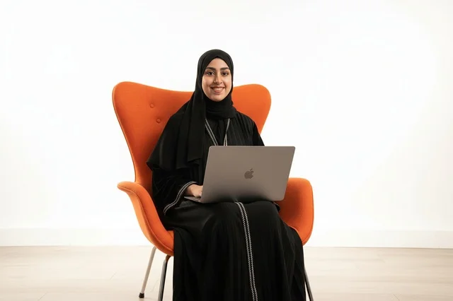 امرأة إماراتية مع لابتوب على كرسي برتقالي خلفية بيضاء
