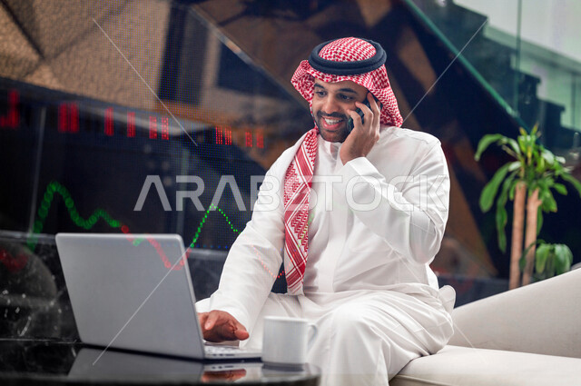 رجل اعمال عربي خليجي سعودي يجلس امام الحاسوب المحمول ويقوم باجراء مكالمة هاتفية لمناقشة اوضاع السوق  ، متابعة مخطط الرسم البياني للنمو الاقتصادي ، تصفح سوق الاوراق المالية ، النجاح في التداول المالي ، مراقبة الاقتصاد والمعلومات المالية ، الاستثمار المالي 