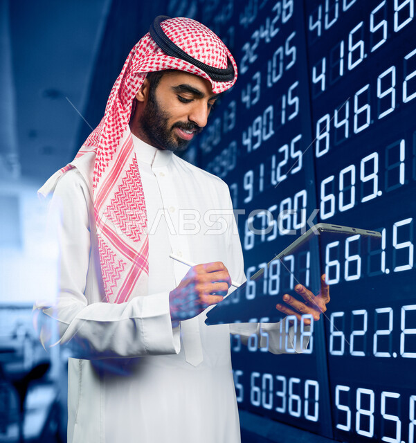 A smiling Saudi Arabian Gulf Arab man using a tablet device, taking notes and information, following the stock market, succeeding in financial trading, monitoring the economy and financial information, financial investment, background of a digital stock m