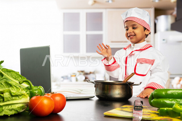 طفل عربي خليجي سعودي ، يرتدي زي الشيف، يقف أمام طاولة عليها مجموعة من الخضراوات الطازجة، إستخدام جهاز الحاسوب المحمول، مشاركة تحضير الوصفات على مواقع التواصل الاجتماعي، إعداد السلطات والأطباق الصحية، مهنة مستقبلية، بث مباشر