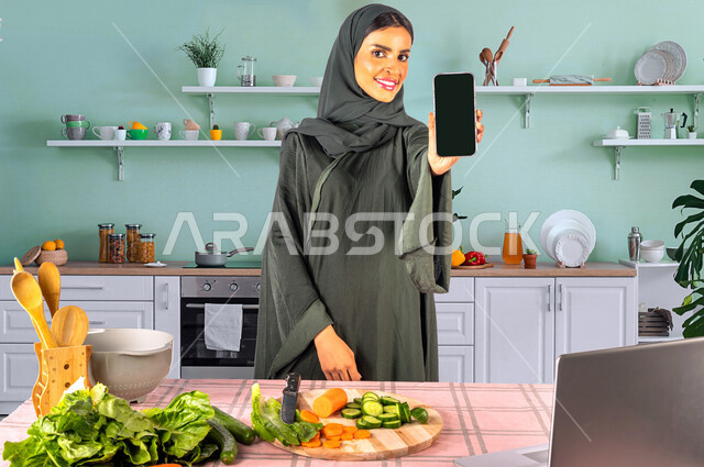 A Saudi Gulf woman, standing in front of a table with a group of fresh vegetables in the kitchen, displaying a black screen, displaying mobile services and applications, using a laptop, sharing preparing recipes on social media, preparing healthy dishes, 