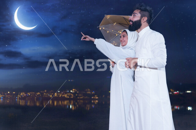 زوجان عربيان سعوديان خليجيان ينظران الى هلال شهر رمضان المبارك, الإشارة الى الهلال, إيماءات وجه تدل على البهجه والسرور بدخول شهر الخير, مشاعر وأجواء رمضانية, مناسبات اسلاميه, إستقبال هلال رمضان