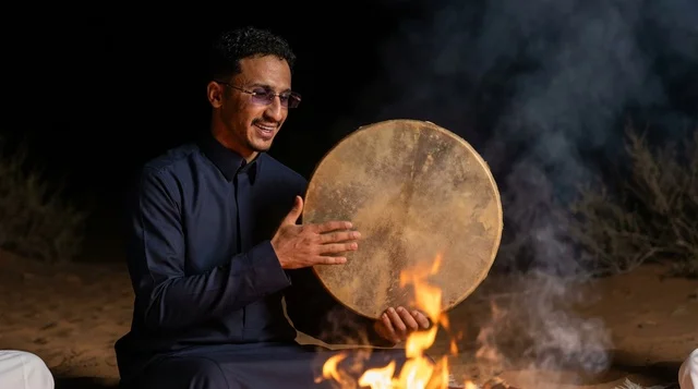 رجل سعودي بزي تقليدي يحمل دفاً تراثياً بجانب نار المخيم