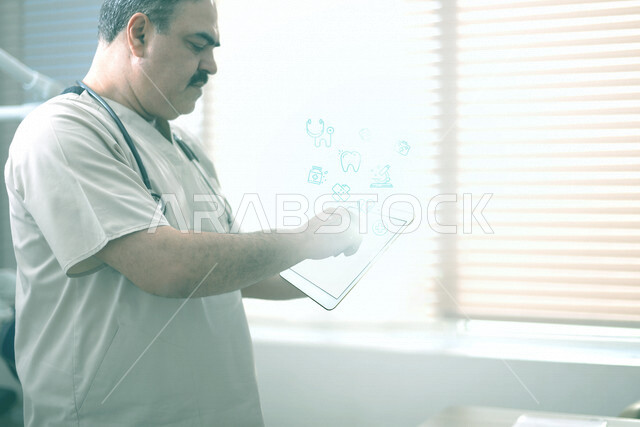 طبيب عربي خليجي سعودي يرتدي السماعة الطبية ويستخدم جهاز التابلت عليه ايقونات هولوجرامية متطايرة ، تقديم الاستشارات الطبية عبر الانترنت ، مراجعة التقارير والسجلات الطبية ، العمل في القطاع الصحي ، مفهوم الطب والرعاية الصحية