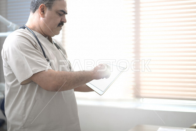 طبيب عربي خليجي سعودي يرتدي السماعة الطبية ويستخدم جهاز التابلت ،تقديم الاستشارات الطبية عبر الانترنت ، مراجعة التقارير والسجلات الطبية ، العمل في القطاع الصحي ، مفهوم الطب والرعاية الصحية