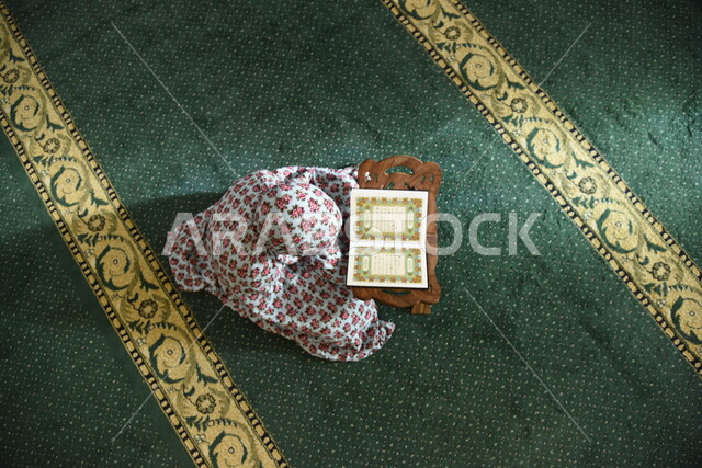 فتاة عربية خليجية ترتدي الزي الإسلامي وتجلس في المسجد وامامها المصحف الشريف على حامل خشبي في المسجد، الصلاه والدعاء في المسجد، العبادة والتقرب الى الله، قراءة وتلاوة القران، التعبد في بيوت الله، ذكر الله، تحفيظ القران الكريم، تفسير القرآن