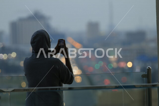 صورة بزاوية من الخلف لرجل سائح يقوم بإلتقاط الصور بإستخدام كاميرا الهاتف المحمول ، خلفية ضبابية ، موهبة وإحترافية فن التصوير ، شوارع المملكة العربية السعودية