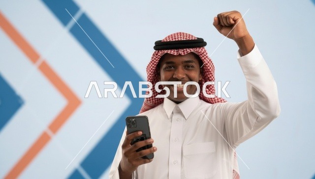 شاب سعودي يمسك هاتف ذكي ويرفع قبضة يده فرحاً بالنجاح