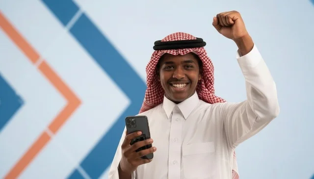 شاب سعودي يمسك هاتف ذكي ويرفع قبضة يده فرحاً بالنجاح