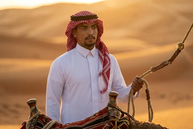 رجل سعودي بالزي التقليدي مع جمل في الصحراء عند الغروب