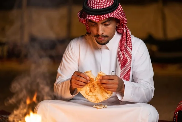 رجل سعودي بالزي التقليدي يأكل الخبز بجانب النار