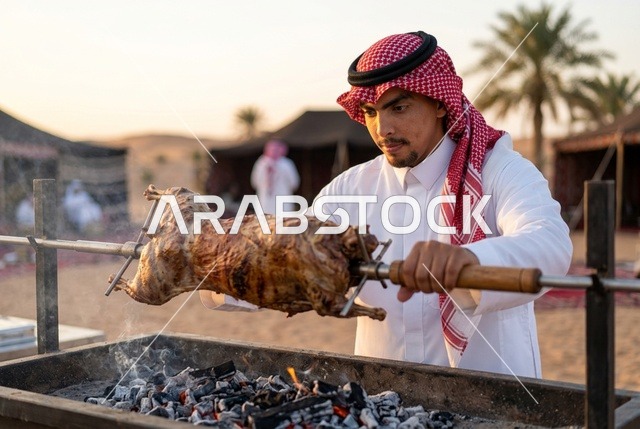 رجل سعودي يرتدي الزي التقليدي يشوي خروفاً في الصحراء