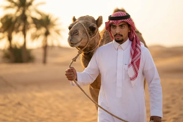 رجل سعودي بالزي التقليدي مع جمل في الصحراء وقت الغروب