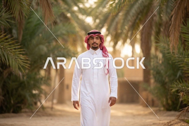شاب سعودي بالزي التقليدي يمشي في واحة النخيل الدافئة