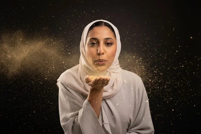 امرأة عربية محجبة تنفخ غبارا ذهبيا لامعا بخلفية سوداء