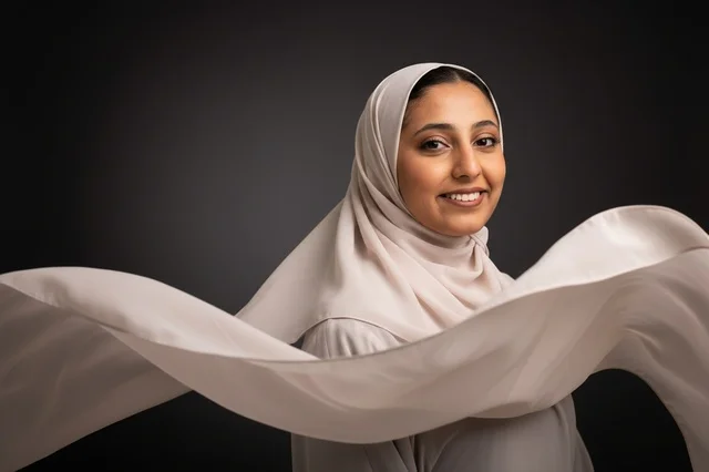 A smiling Arab woman wearing a flowing hijab against a black background.