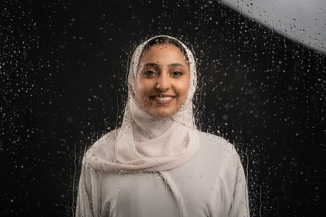 امرأة عربية مبتسمة بالحجاب خلف زجاج عليه قطرات مطر