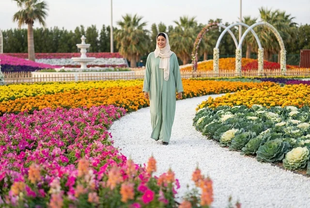 امرأة عربية بعباءة خضراء تمشي في حديقة زهور ملونة