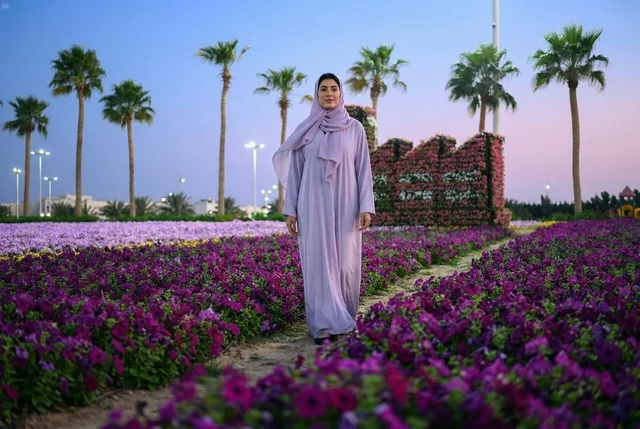 امرأة عربية بالعباءة تقف في حديقة زهور ملونة وقت الغسق