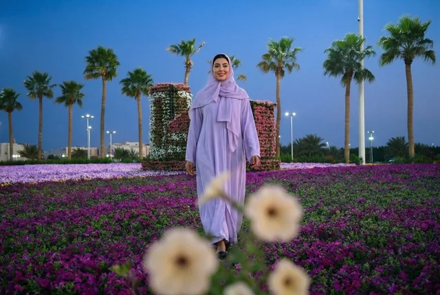 امرأة عربية بعباءة بنفسجية تمشي في حديقة زهور بالمساء