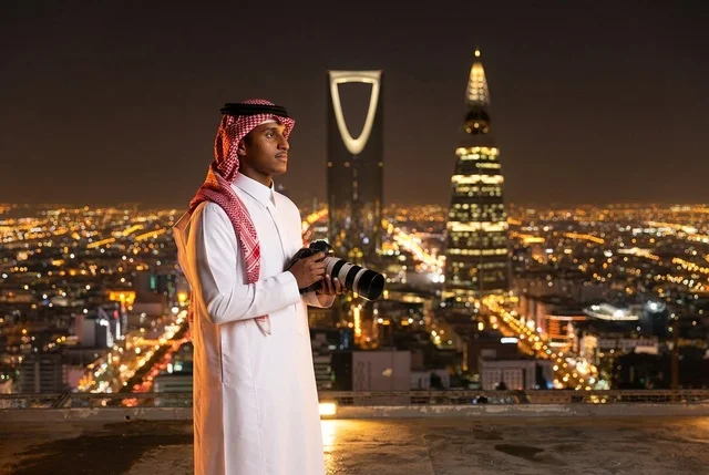 مصور سعودي يحمل كاميرا أمام أفق مدينة الرياض ليلا