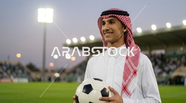 شاب سعودي بالثوب التقليدي يحمل كرة قدم بالملعب