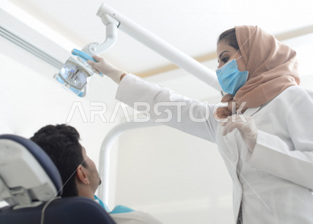 طبيبة اسنان عربية خليجية سعودية ترتدي المعطف الطبي والكمامة الطبية ، استخدام اجهزة تقنية طبية لفحص وتشخيص اسنان المريض ، معالجة التسوس وتركيب الحشوات ،تقديم الخدمات الصحية، طب وجراحة الفم والاسنان