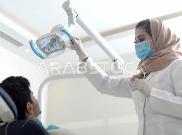 طبيبة اسنان عربية خليجية سعودية ترتدي المعطف الطبي والكمامة الطبية ، استخدام اجهزة تقنية طبية لفحص وتشخيص اسنان المريض ، معالجة التسوس وتركيب الحشوات ،تقديم الخدمات الصحية، طب وجراحة الفم والاسنان