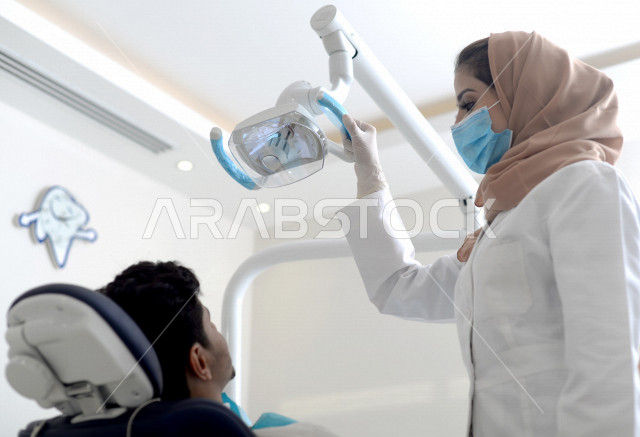 طبيبة اسنان عربية خليجية سعودية ترتدي المعطف الطبي والكمامة الطبية ، استخدام اجهزة تقنية طبية لفحص وتشخيص اسنان المريض ، معالجة التسوس وتركيب الحشوات ،تقديم الخدمات الصحية، طب وجراحة الفم والاسنان