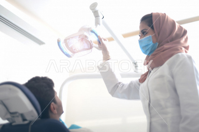 طبيبة اسنان عربية خليجية سعودية ترتدي المعطف الطبي والكمامة الطبية ، استخدام اجهزة تقنية طبية لفحص وتشخيص اسنان المريض ، معالجة التسوس وتركيب الحشوات ،تقديم الخدمات الصحية، طب وجراحة الفم والاسنان