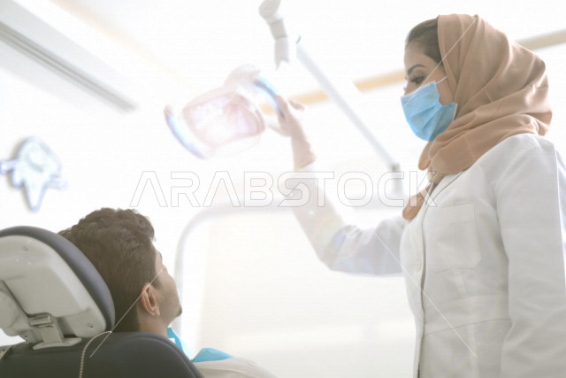 طبيبة اسنان عربية خليجية سعودية ترتدي المعطف الطبي والكمامة الطبية ، استخدام اجهزة تقنية طبية لفحص وتشخيص اسنان المريض ، معالجة التسوس وتركيب الحشوات ،تقديم الخدمات الصحية، طب وجراحة الفم والاسنان
