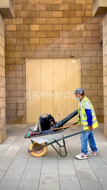 صورة لعامل النظافة ( مهندس الصحة ) يرتدي الزي الخاص بالعمل يجرعربة التنظيف التي تحتوي على أدوات النظافة الخاصة بالعمل ، تنظيف الأبنية و الطرق و الأرصفة ، الاهتمام بنظافة البيئة و رونقها