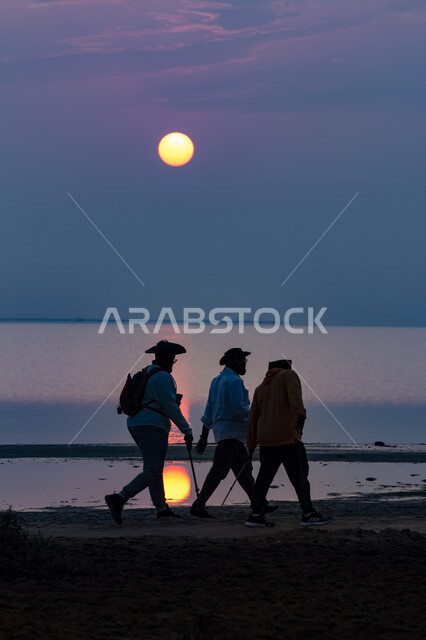 صورة جمالية لمجموعة من السائحين يسيرون على شاطئ البحر وقت غروب الشمس ، إطلالة رائعة للشمس في السماء و انعكاسها على مياه البحر ، جولة سياحية طبيعية ترفيهية 