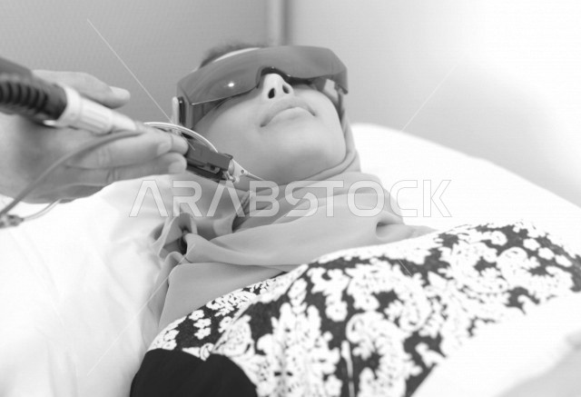 طبيب عربي خليجي سعودي يستخدم جهاز الليزر على وجه سيدة خليجية سعوية ترتدي النظارة الواقية ، اجهزة تقنية طبية حديثة ، تقديم الخدمات الصحية، طب الجلدية ومجال العناية بالبشرة